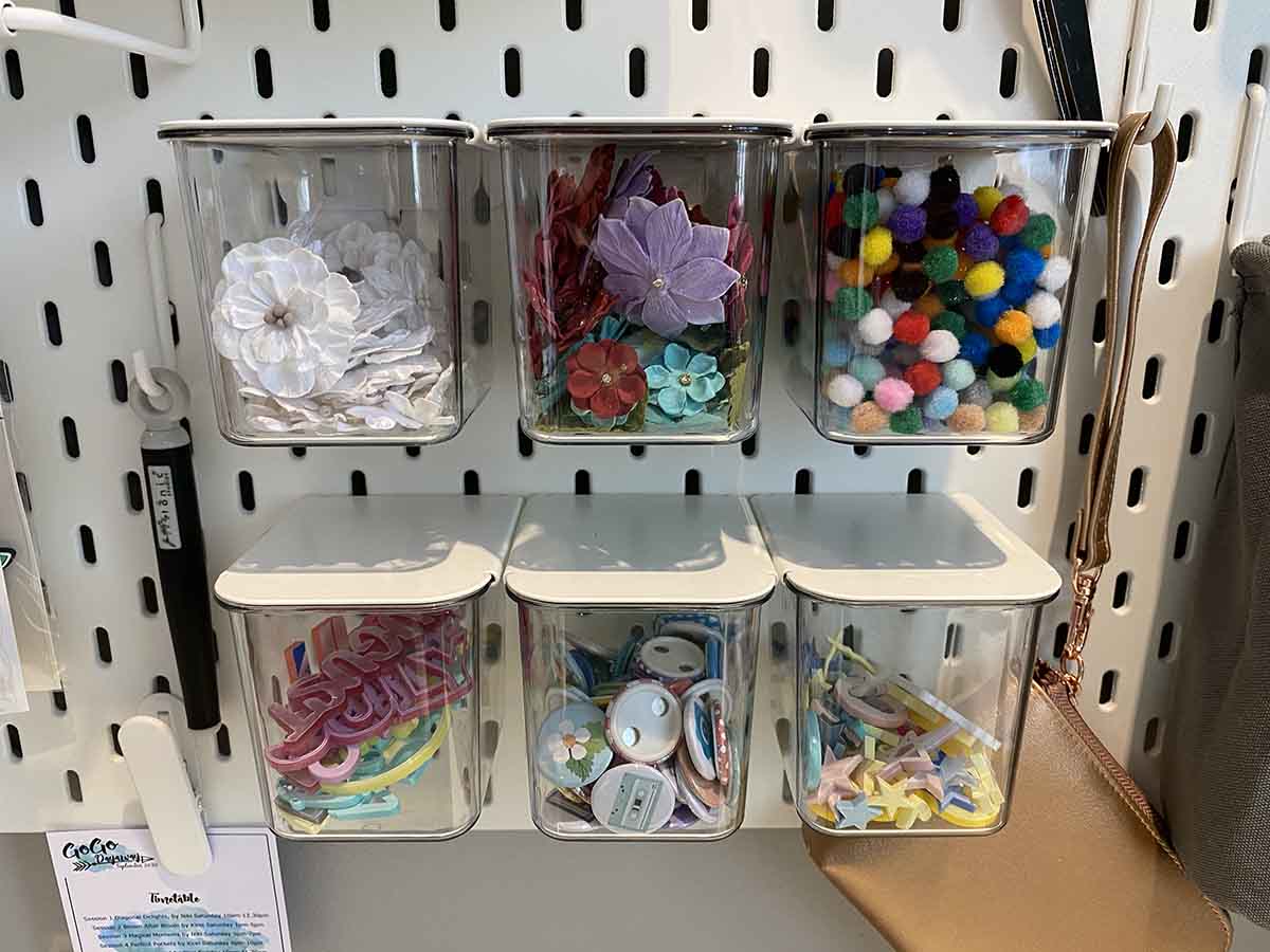 Ikea Skadis containers with lids on pegboard in craftroom filled with buttons, flowers and scrapbooking embellishments.