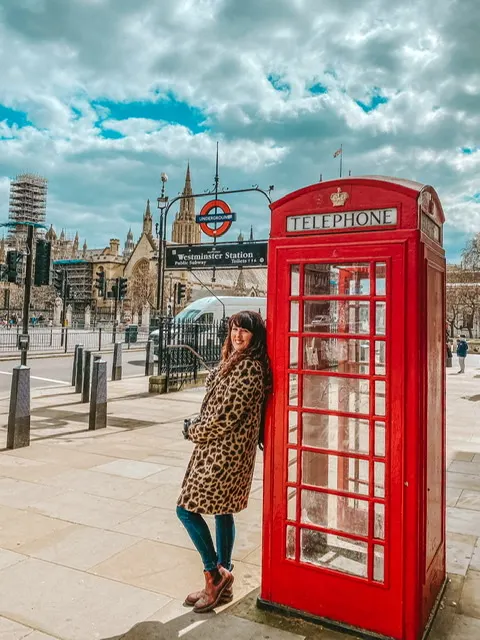 London phone box outside the House of Parliament. Woman leaning on box with Westminster station behind 